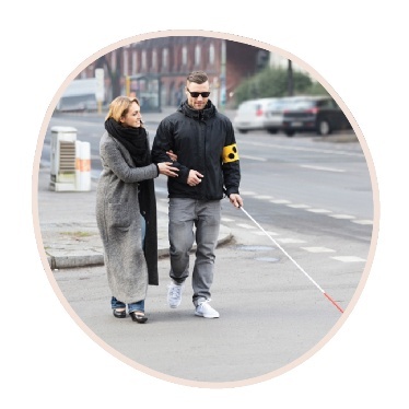 A person disability using a white cane. Someone else is walking with them, holding their arm.