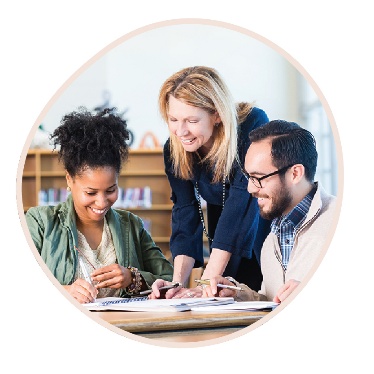A person standing and leaning in to help 2 people sitting at a table with their work.